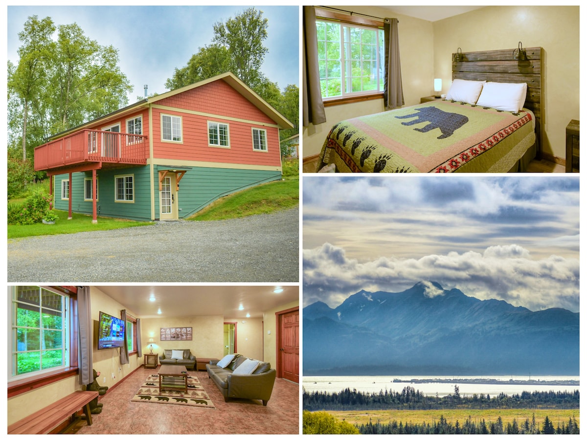 The exterior of Bear Creek Cottage features a two-story structure with a mix of green and red siding, surrounded by greenery. A gravel driveway leads to the home, where large windows allow natural light into the living space. A scenic view of mountains and Kachemak Bay is captured in the image.
