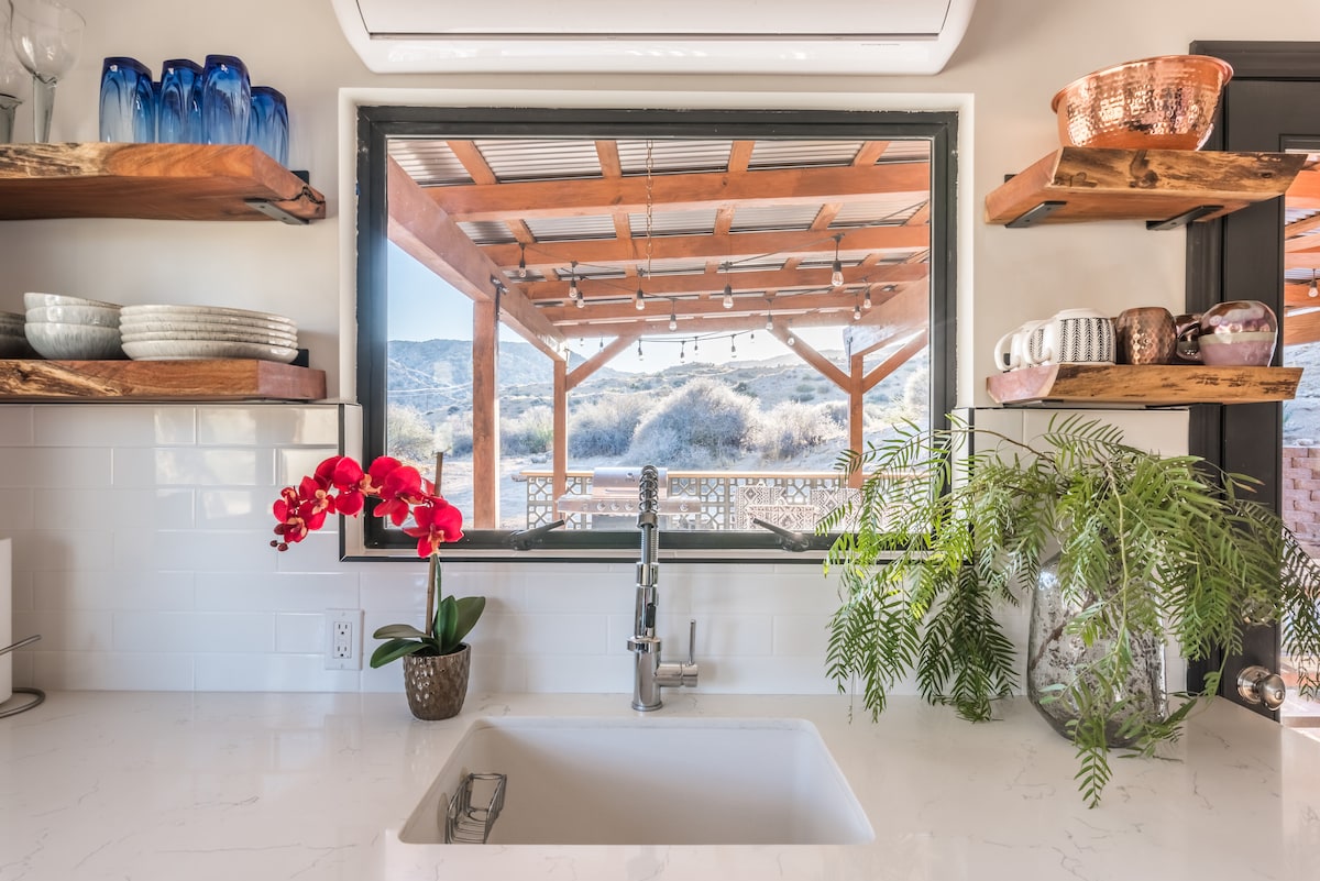 A modern kitchen sink is positioned beneath a large window showcasing desert views. Wooden shelves display a variety of dishes, glassware, and plants. Natural light illuminates the clean countertops, creating a bright and airy atmosphere.