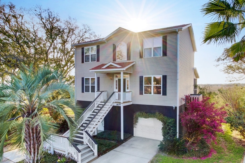 The elevated home features a welcoming entrance with a front staircase leading to a spacious upper level. Lush palm trees and vibrant flowering shrubs are present in the landscaped yard, illuminated by sunlight filtering through the trees.