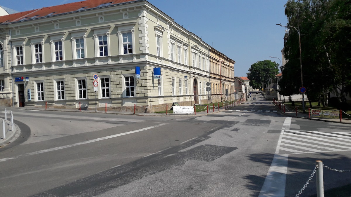A view of a street intersection features a historic building with large windows and classical architecture. Clear signage indicates the direction of traffic, and landscaping with trees is visible in the background. Pedestrian markings are present on the road, enhancing navigability.