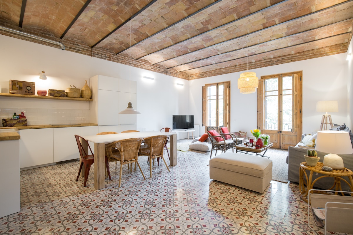 An open living area features high ceilings with exposed beams and a patterned tiled floor. A wooden dining table is surrounded by wicker chairs. A comfortable sectional sofa with colorful cushions is arranged near a window, offering natural light and views of the outside.