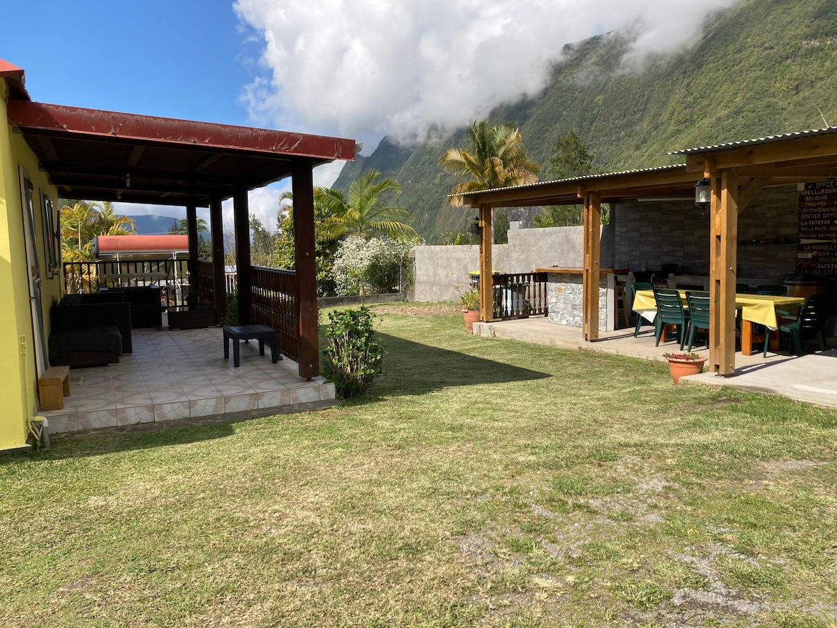 An outdoor area is presented with a spacious lawn bordered by greenery. A covered patio features seating arrangements, while a nearby outdoor kitchen displays dining tables under a shaded structure, all set against a backdrop of distant mountains and a clear blue sky.