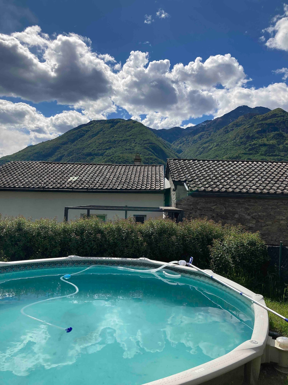 A private pool is featured in the foreground, surrounded by a lush green garden. In the background, majestic mountains are framed by a clear blue sky scattered with clouds. Nearby structures are visible, presenting a serene residential setting.