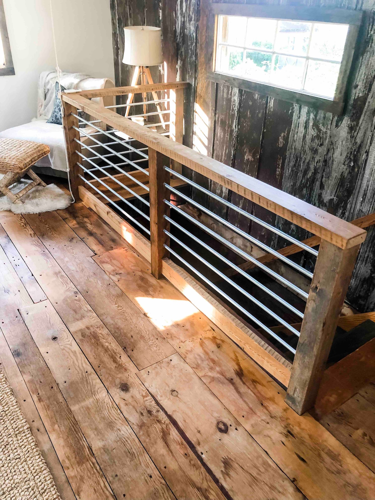 A wooden railing is visible, featuring a modern design with horizontal metal slats. Sunlight filters through a nearby window, illuminating the rustic wood flooring below. In the background, a cozy seating area can be seen, creating an inviting atmosphere.