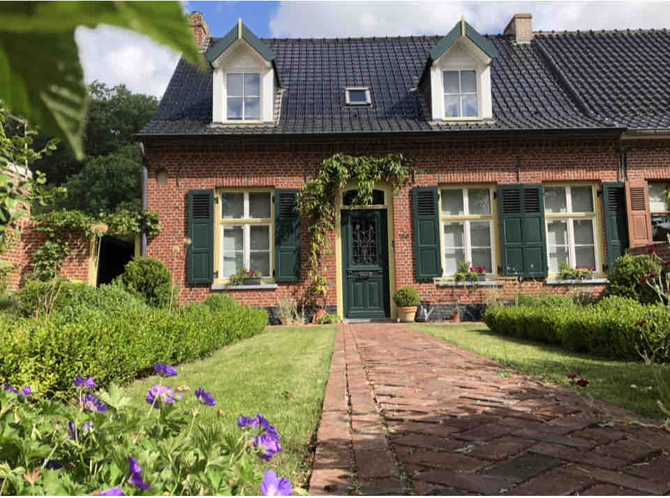 A charming brick house is framed by neatly trimmed hedges and blooming flowers. Green shutters adorn the windows, while a green door welcomes visitors. A well-maintained pathway leads from a garden filled with purple blossoms to the entrance, inviting exploration of the property.