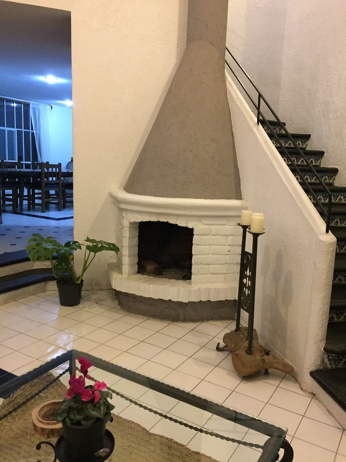 An inviting living space is highlighted by a white brick fireplace, adding a cozy touch. A potted plant is positioned nearby, and candles are placed on a wrought iron stand. Stairs lead to the upper level, with a large dining area visible in the background.