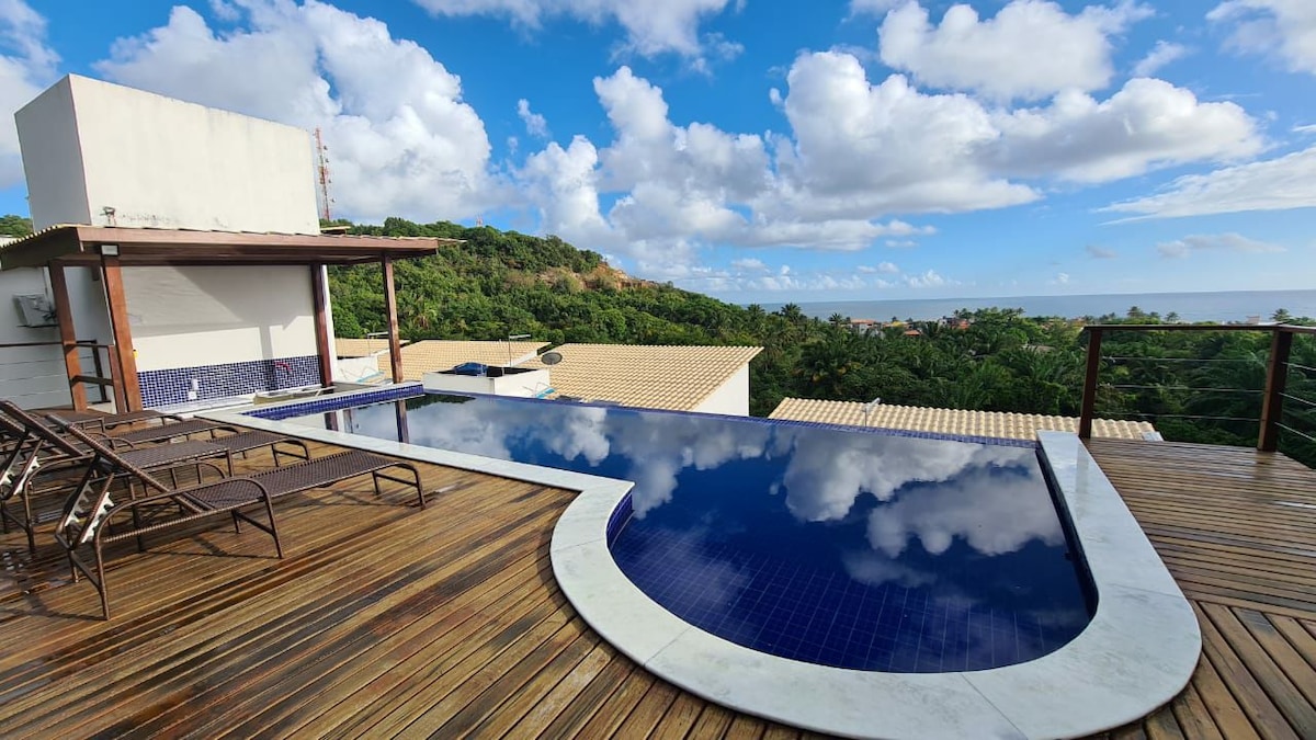 An infinity pool is set on a wooden deck, reflecting the sky and surrounding greenery. Lounge chairs are arranged nearby, providing space for relaxation. The tranquil view includes hills in the distance, enhancing the serene atmosphere of the area.