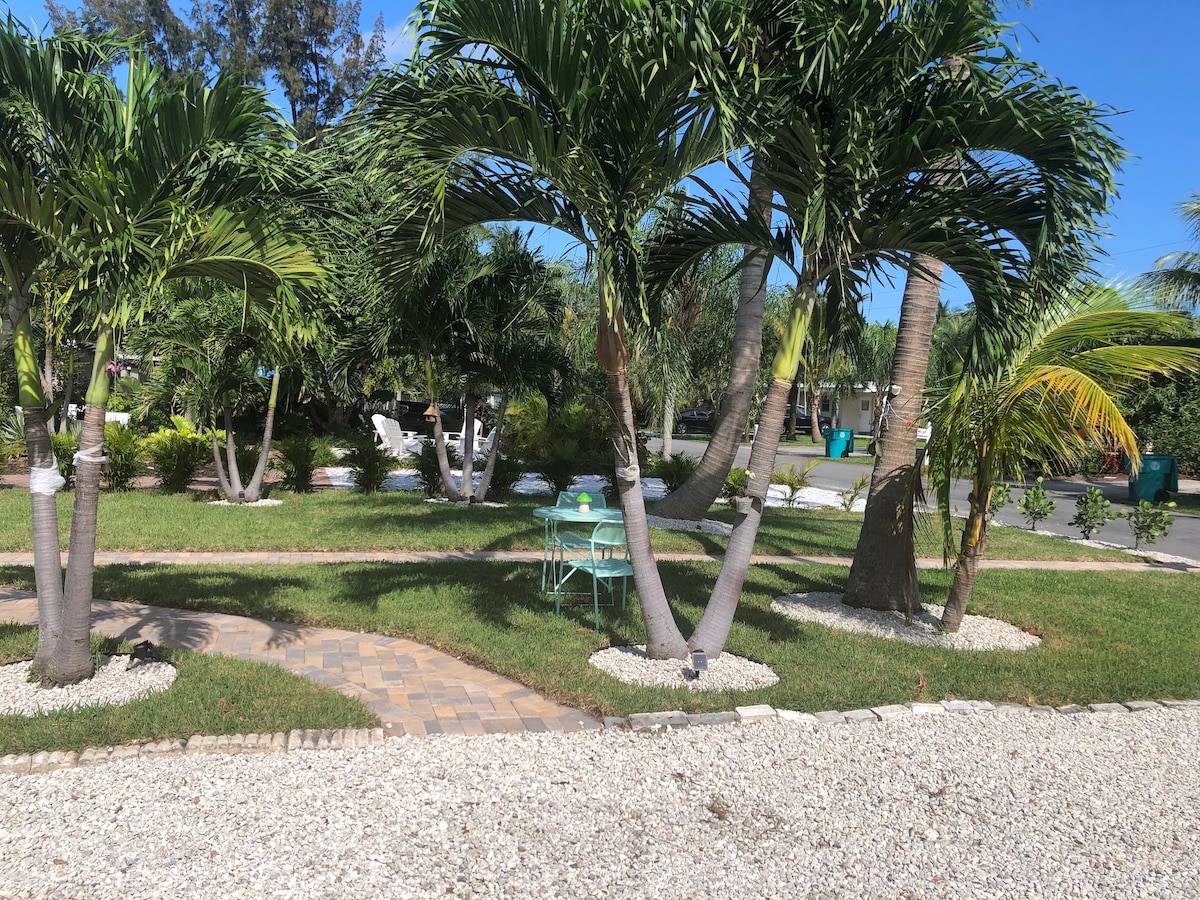 A landscaped yard features lush palm trees and tropical plants, creating a serene outdoor space. A stone pathway winds through the grass, leading to a small seating area with a round table and chairs, set under the shade of the trees.