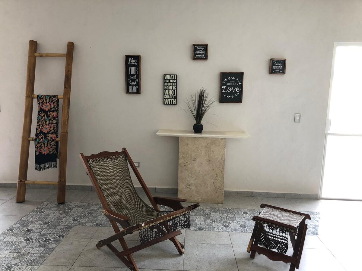 A spacious living area features a wooden ladder displaying a colorful textile. Framed artwork is mounted on light-colored walls, providing a welcoming ambiance. A comfortable chair and matching footrest are positioned on tiled flooring, while natural light enters through a nearby door.