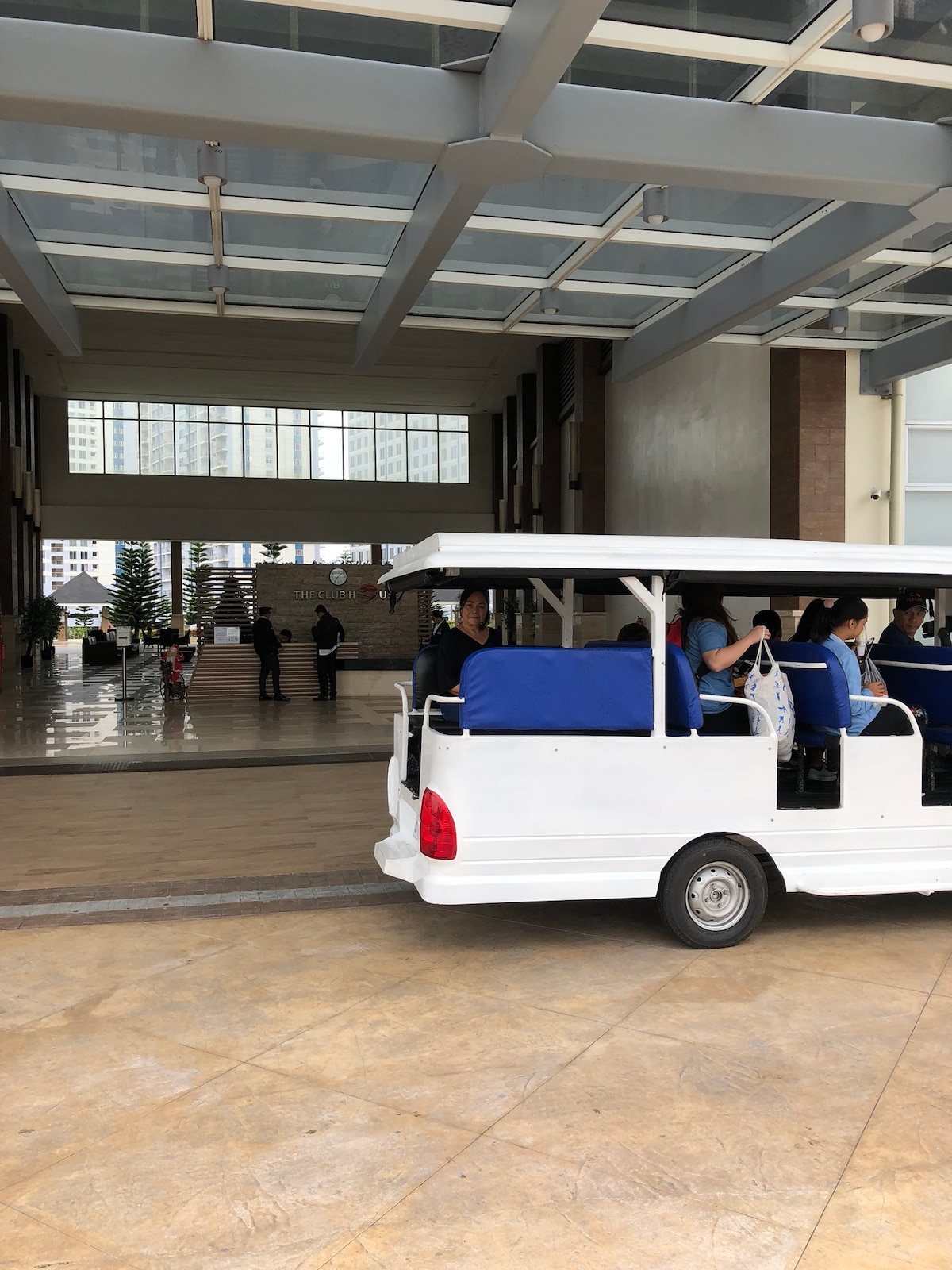A shuttle vehicle, designed for guest transport, is parked at the entrance of a modern building, revealing a spacious lobby with high ceilings and large windows. Smooth flooring leads to a staircase in the background, while green plants are visible, adding to the welcoming environment.