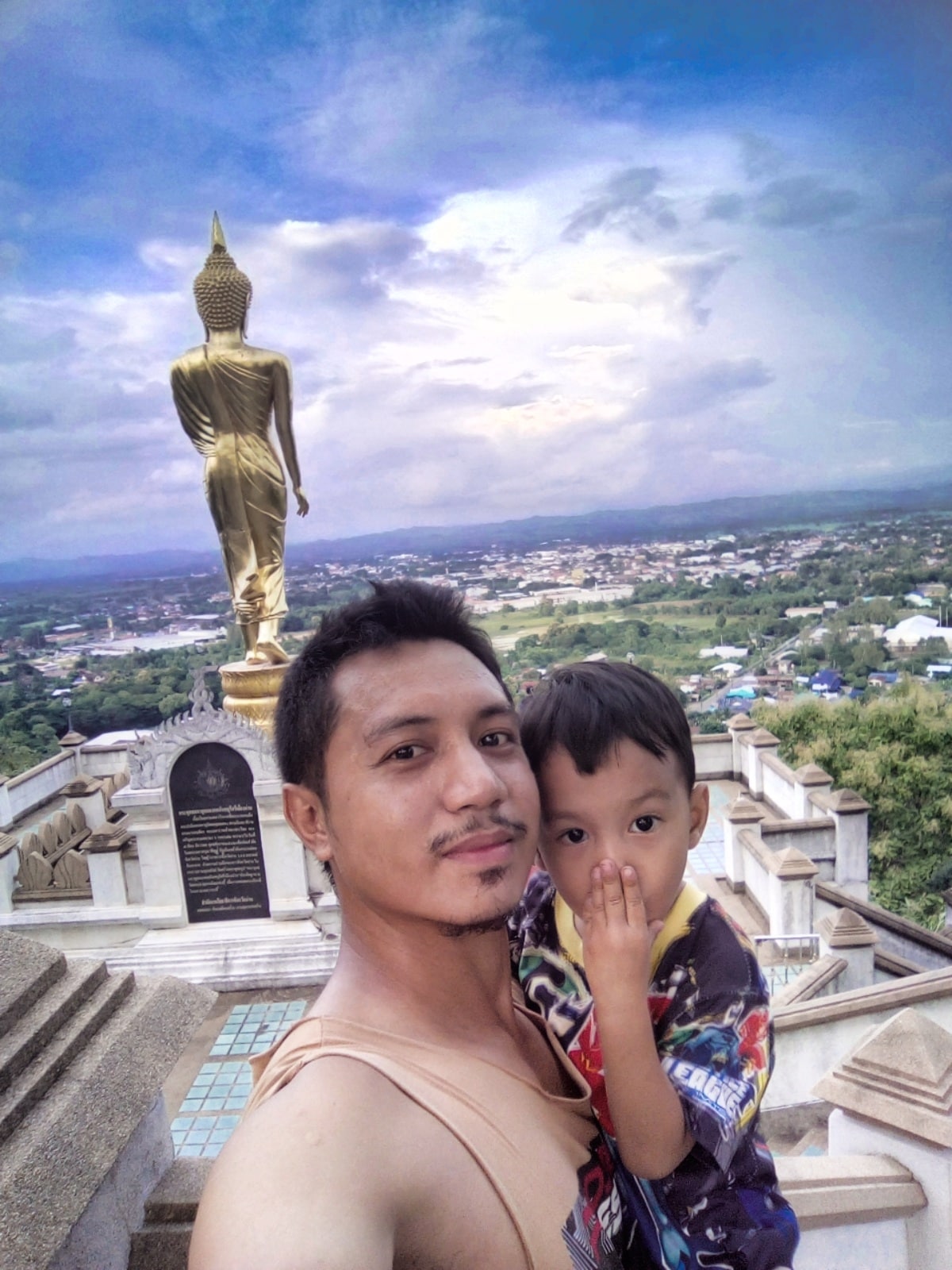 A towering Buddha statue is visible in the background, overlooking a scenic landscape. Below, the valley and town extend under a blend of clouds and blue sky. The image captures the serene atmosphere of this cultural site, contributing to a sense of tranquility and elevation.