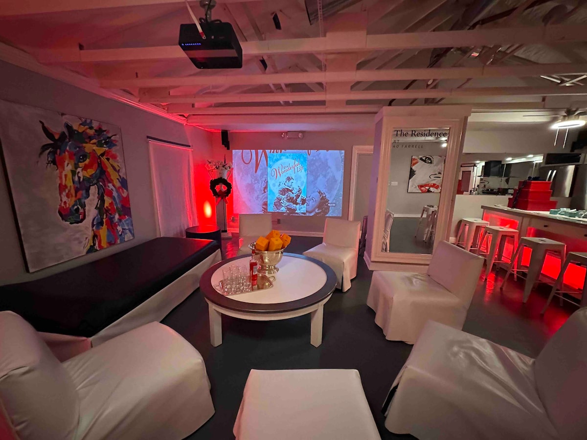 A gathering room is featured, showcasing a large projection screen displaying colorful imagery. Soft white seating surrounds a circular coffee table, adorned with a crystal bowl of fruit. Ambient red lighting enhances the modern feel, complemented by vibrant artwork on the walls.