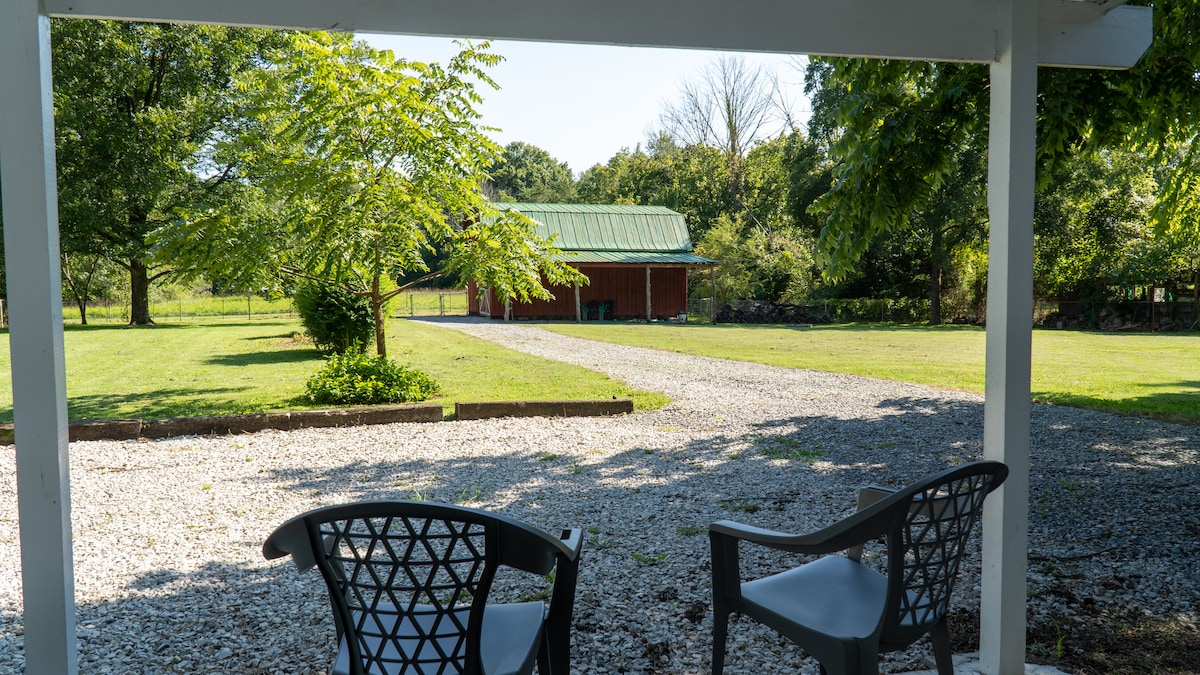 Two chairs are positioned under a covered area, facing an expansive grassy yard. A gravel path leads towards a barn-style structure with a green metal roof, framed by trees and open countryside, creating a serene outdoor setting.