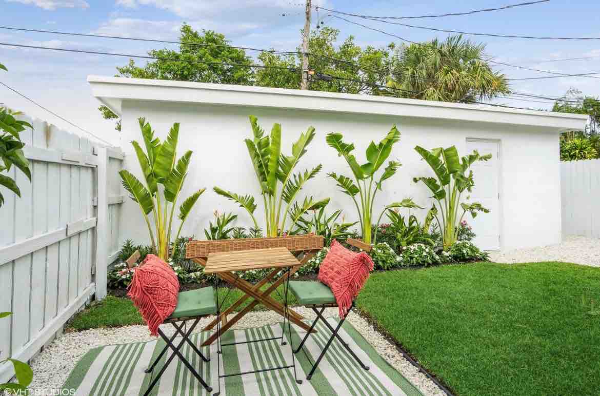An outdoor patio area is presented with a small folding table and two green chairs accented by coral cushions. Lush tropical plants line the background, complementing the green grass and patterned outdoor rug beneath the table, creating an inviting space for relaxation or dining.
