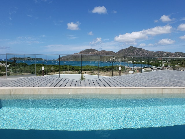 A serene view of the azure swimming pool merges with the daylight sky, showcasing the gentle waves of the bay. In the background, lush hills and distant boats are visible, framed by a modern railing that enhances the outdoor space.