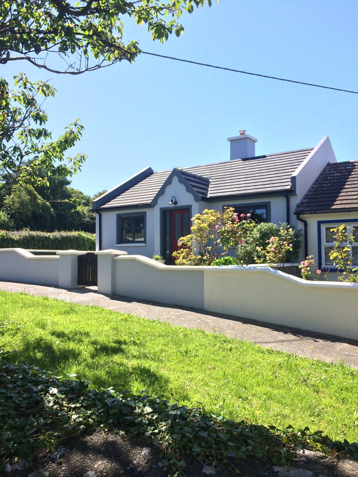 A charming detached cottage is depicted with a vibrant red door framed by well-maintained flowerbeds. Lush greenery surrounds the property, and the clear blue sky suggests a bright day. A paved pathway leads to the entrance, emphasizing the homey atmosphere.