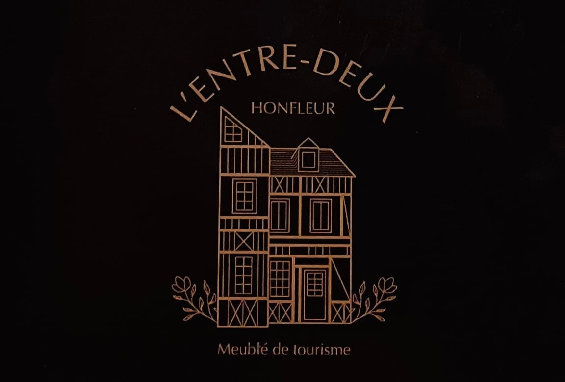 The image displays a stylized illustration of the building 'L'Entre-Deux' in Honfleur. The design features an elegant façade with distinct architectural elements, including windows and a peaked roof, framed by delicate floral motifs below the name of the property.