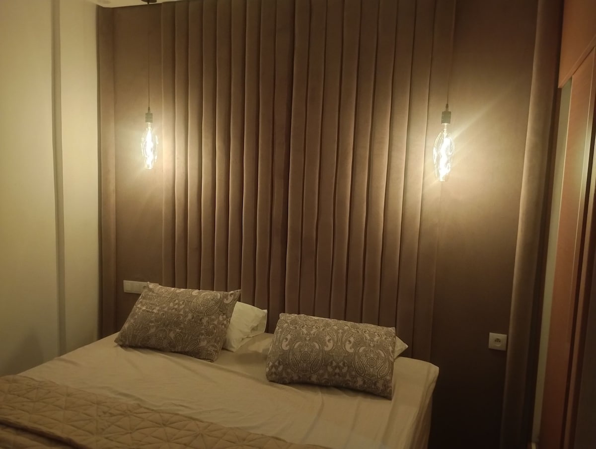 A relaxing sleeping area features a soft, taupe tufted headboard with modern lighting fixtures on either side. Two decorative pillows are placed on a spacious bed, while a light coverlet is neatly arranged at the foot.