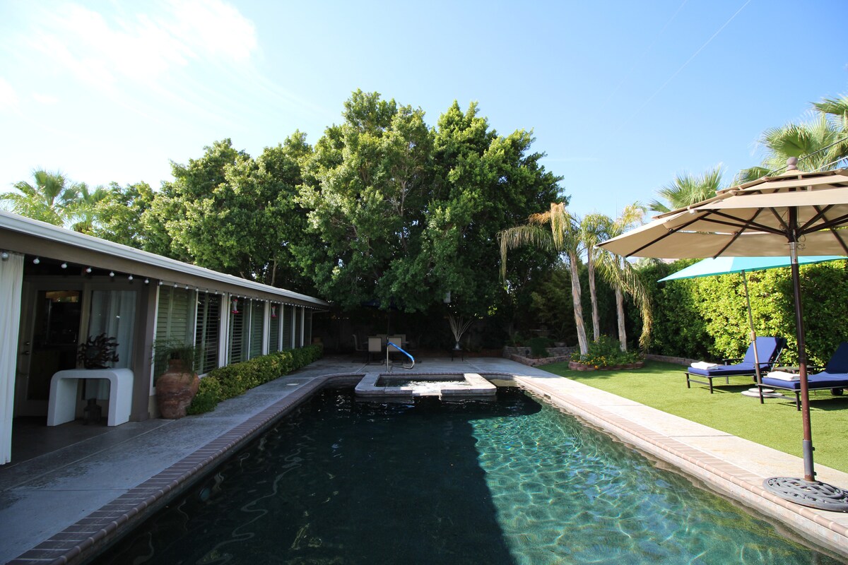 A lively outdoor space features a blue-tiled pool surrounded by lush greenery and tall palm trees. A stone patio offers lounging options with a sun umbrella and chaise lounges, adding to the serene atmosphere. The sun shines brightly in a clear blue sky.
