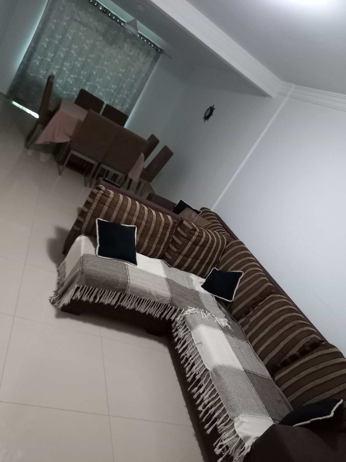 A spacious living area features a brown striped sectional sofa adorned with decorative cushions. A cozy throw blanket drapes over the sofa. In the background, a dining table with upholstered chairs is visible, illuminated by natural light filtering through sheer curtains.