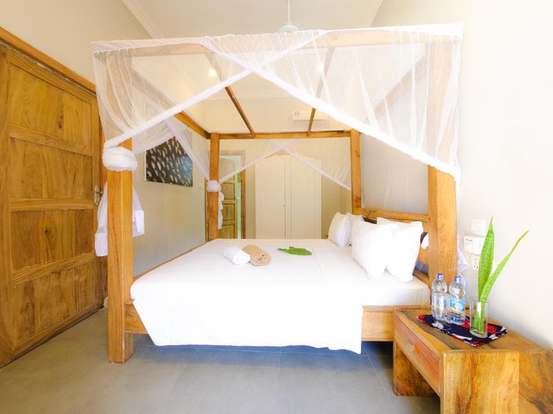 A spacious bed with a wooden frame is adorned with a white canopy and crisp linens. Two bottles of water are placed on a side table, accompanied by a small green plant. Natural light fills the room, highlighting the warm wooden door and accents.