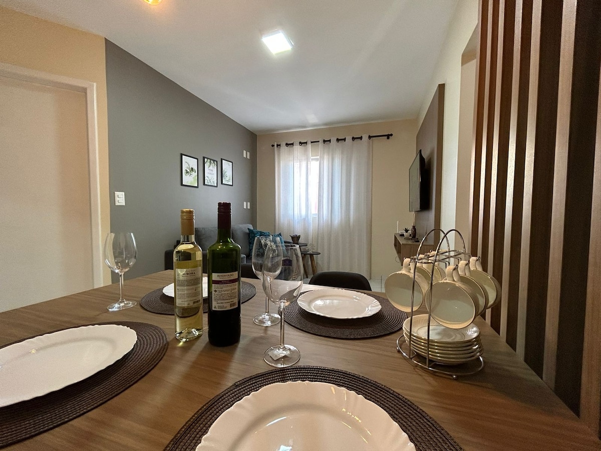 A dining table is set with four plates, wine glasses, and two bottles of wine. In the background, a cozy living area is visible, featuring a sofa and a wall-mounted television. Natural light filters through sheer curtains, enhancing the comfortable ambiance.