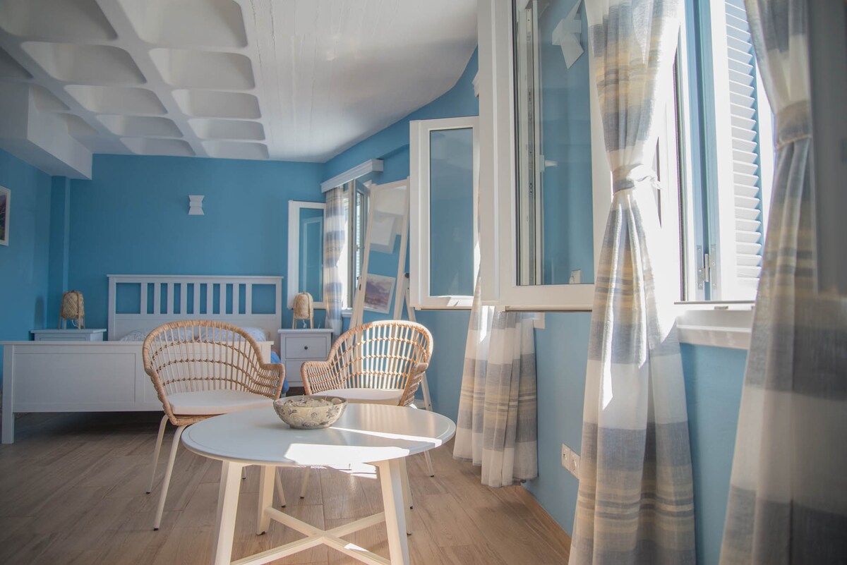 A bright living area features light blue walls and large windows that allow ample natural light. Two rattan chairs and a round table are positioned on the wooden floor, with soft drapes framing the windows. Modern decor adds to the welcoming ambiance.