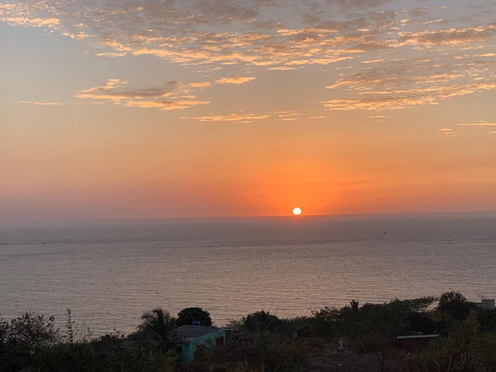 A vibrant sunset is captured over the calm sea, with warm orange and yellow hues reflecting on the water's surface. The sky is adorned with soft clouds, creating a serene atmosphere. The landscape includes gentle hills and sparse vegetation in the foreground.