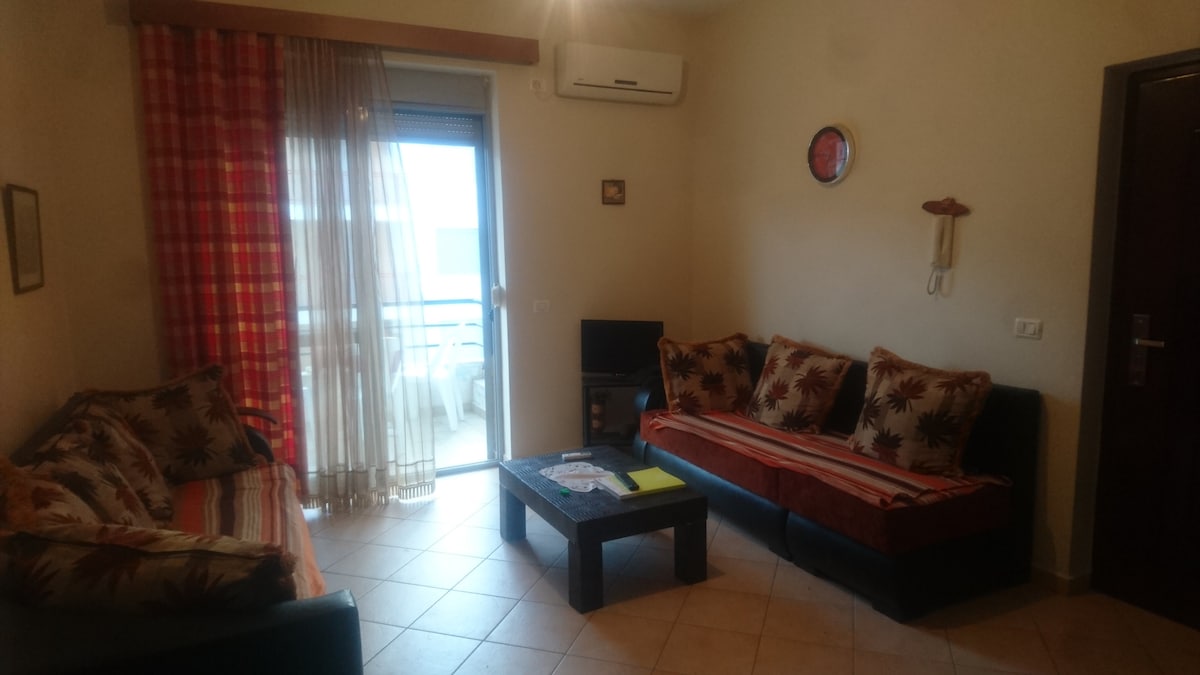 A cozy living area features two sofas adorned with patterned cushions, a coffee table positioned at the center, and a flat-screen TV nearby. Natural light enters through large glass doors, leading to a balcony. A partially decorated wall adds a touch of character.