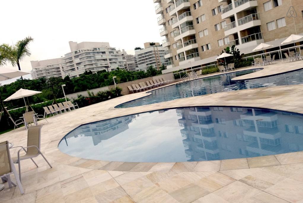 A resort-style pool area features a curved, clear water pool surrounded by a spacious sun deck. Umbrellas shade several lounge chairs positioned around the pool. Lush greenery is visible in the background, with tall condominium buildings framing the scene.