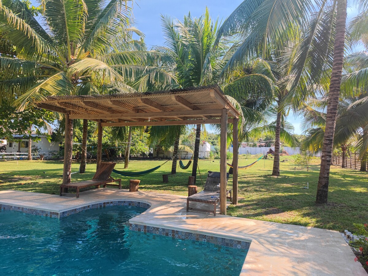 A private pool is surrounded by lush greenery and palm trees. A shaded area with wooden furniture and hammocks offers a relaxing spot. Sunlight reflects on the water, inviting moments of leisure and comfort.