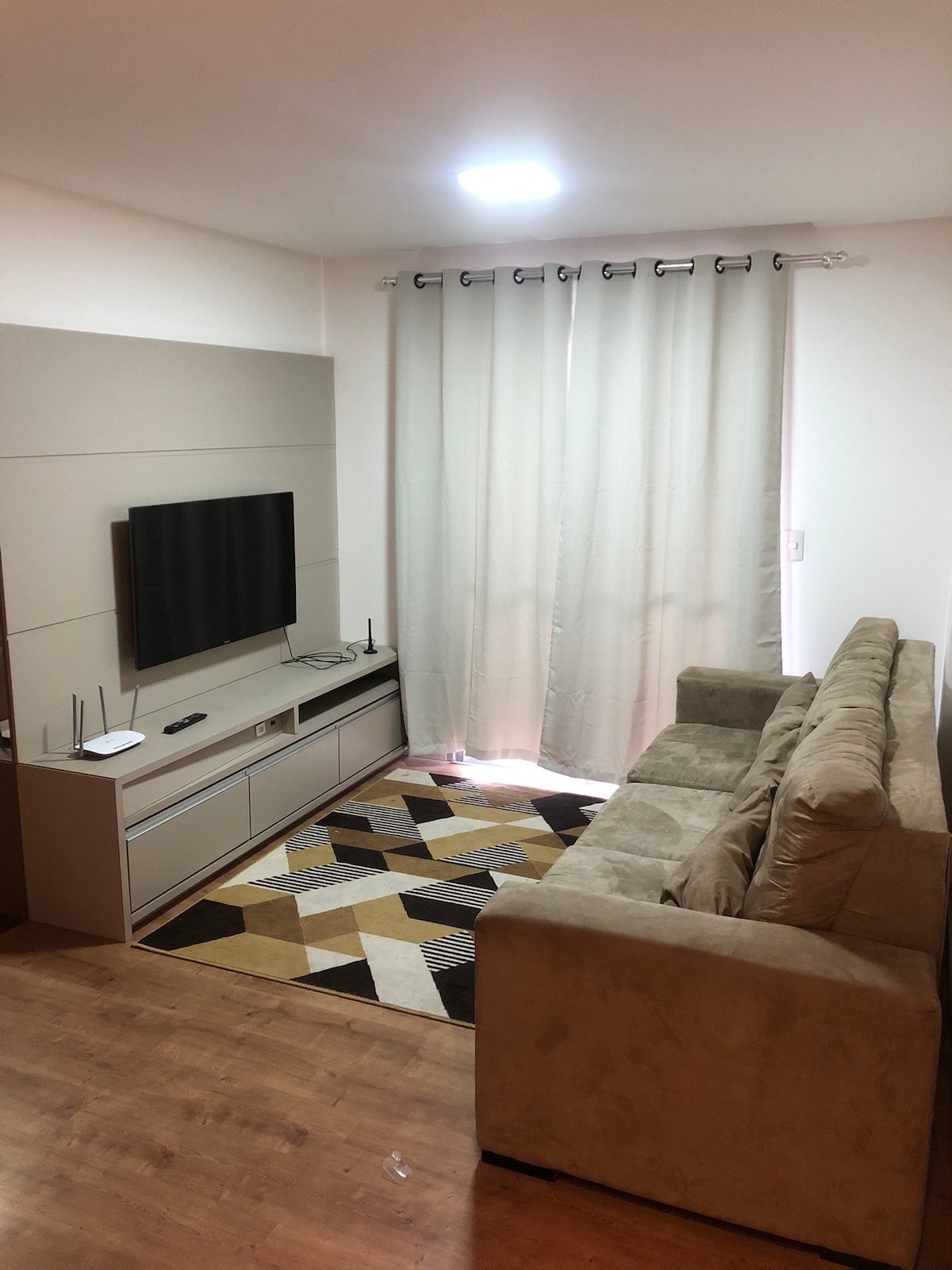A modest living area features a beige sofa arranged opposite a wall-mounted television. A light-colored entertainment center is positioned beneath the screen. Natural light enters through a set of sheer curtains, and a geometric rug adds visual interest to the hardwood floor.