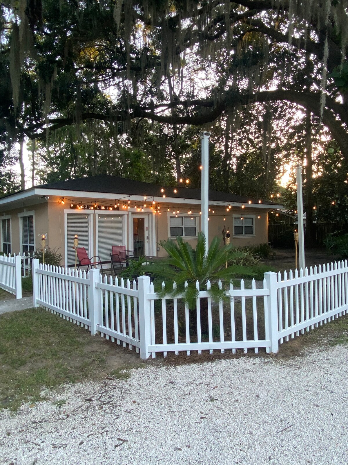A charming single-story home is surrounded by a white picket fence, featuring string lights that softly illuminate the exterior. Lush greenery and palm plants are visible in the front yard, creating a welcoming ambiance against the backdrop of mature trees.