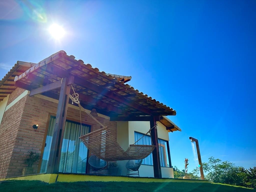 A shaded outdoor area features a cozy hammock suspended between two posts, inviting relaxation. The sun radiates brightly in the clear blue sky above. Surrounding vegetation adds a touch of greenery, complementing the rustic architecture of the property.