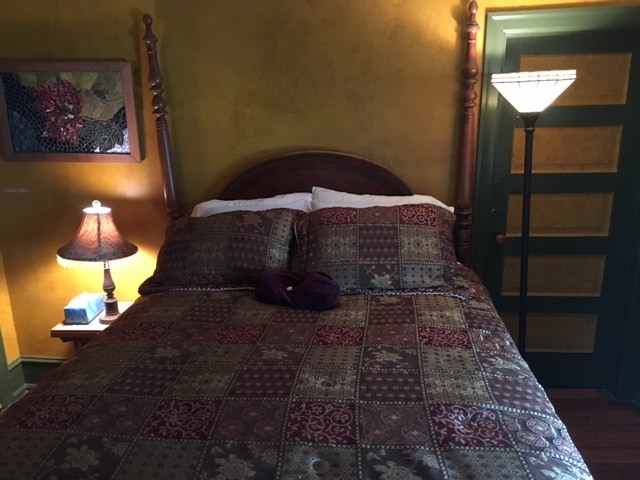 A sleeping area features a wooden bed with intricately carved posts, dressed in a richly patterned comforter. Bedside tables are positioned on either side, each holding a lamp that casts a warm light, while a decorative piece adorns the wall above. A green door adds a contrasting touch.