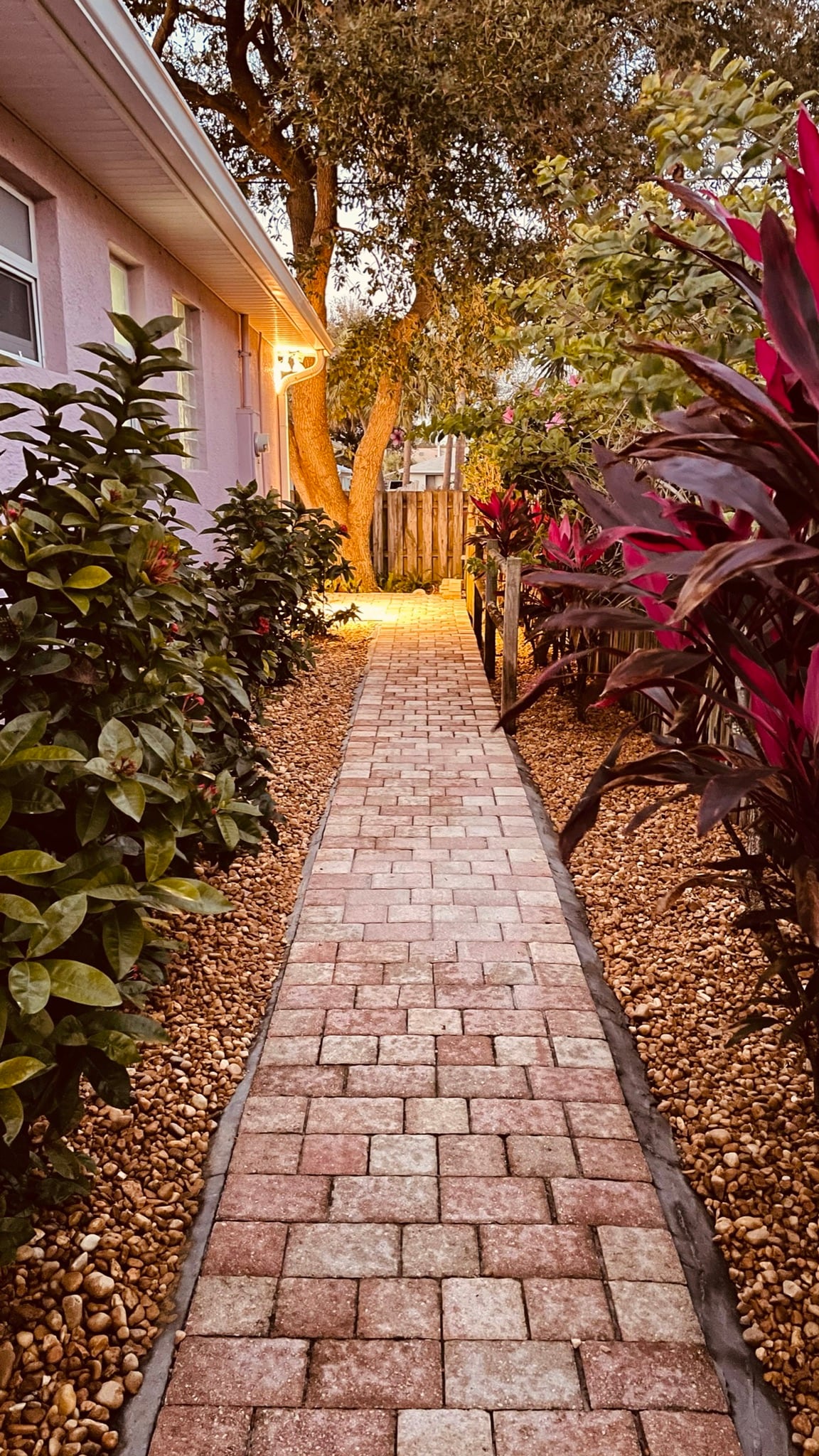 A well-maintained pathway made of patterned brick leads between lush, flowering plants. Soft lighting from above casts a warm glow along the walkway, inviting guests to explore the front entrance of the property.