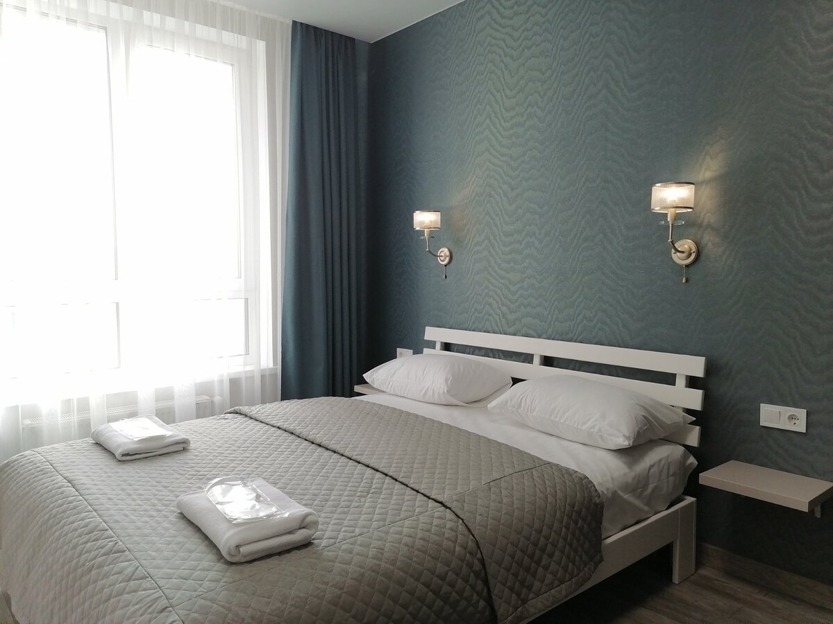 A cozy sleeping area features a spacious bed, dressed with a light grey quilt and topped with two white pillows. Soft lighting is provided by sconces on either side. Natural light fills the room through a large window, complemented by sheer curtains.