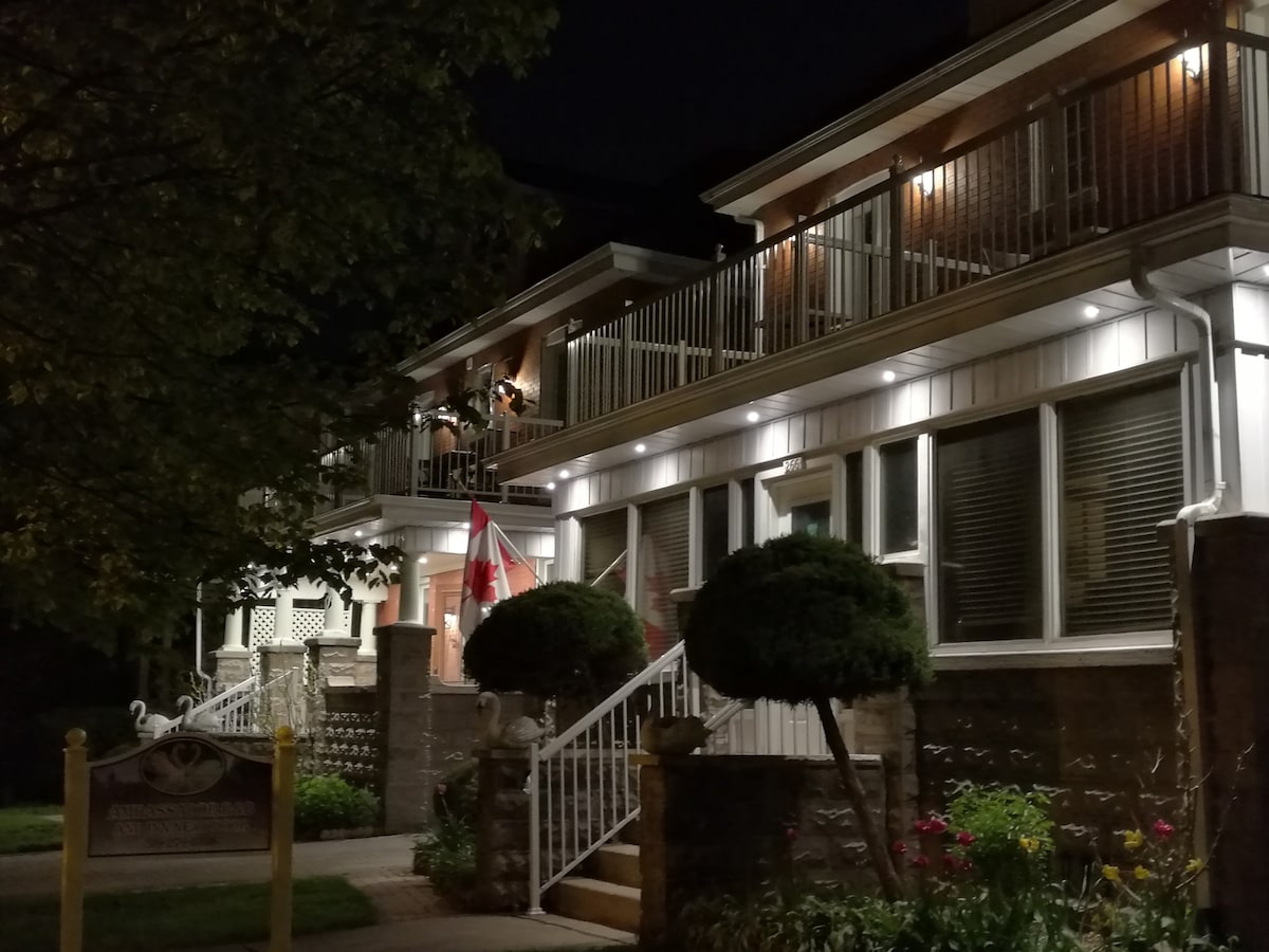 The exterior of a Victorian house is illuminated at night, featuring decorative lighting that highlights the architectural details. A Canadian flag is placed prominently at the entrance, while well-maintained shrubbery and flowers enhance the welcoming appearance of the property.