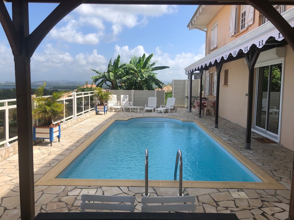 A private swimming pool is framed by a stone deck, with two poolside chairs and vibrant greenery nearby. The covered terrace provides shaded seating and views of the surrounding landscape. Light blue water reflects the sky, creating a calm ambiance for relaxation.
