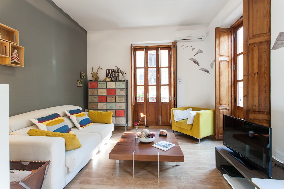 A bright living area features a white sofa adorned with colorful cushions and a cozy yellow armchair. A wooden coffee table rests on light hardwood flooring, while a flat-screen TV sits on a modern stand. Natural light streams through large windows framed in wooden shutters.
