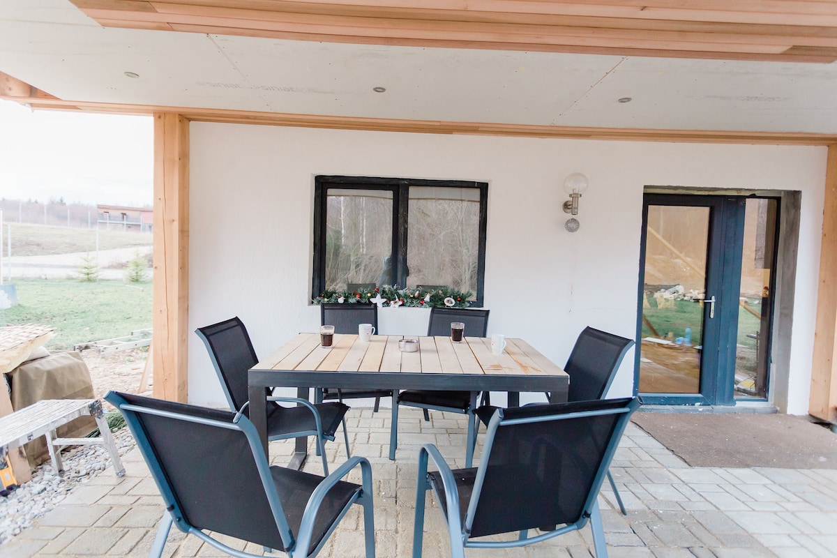 An outdoor patio area is equipped with a large wooden table surrounded by four black chairs. The space features a white exterior wall and a door with a glass panel leading inside. Decorative flower boxes are situated beneath the windows.