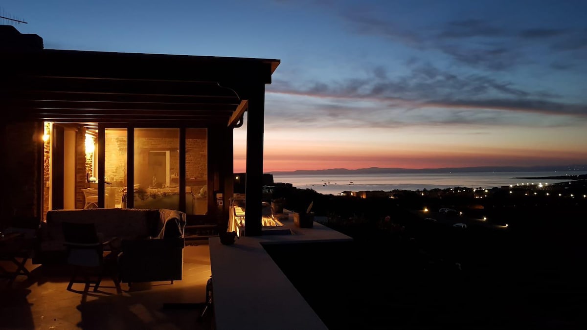 The image captures a two-story home at dusk, with warm lighting illuminating the interior. A covered terrace presents views of a vibrant sunset over the ocean, with soft clouds enhancing the evening sky. The landscape features distant lights along the coastline.