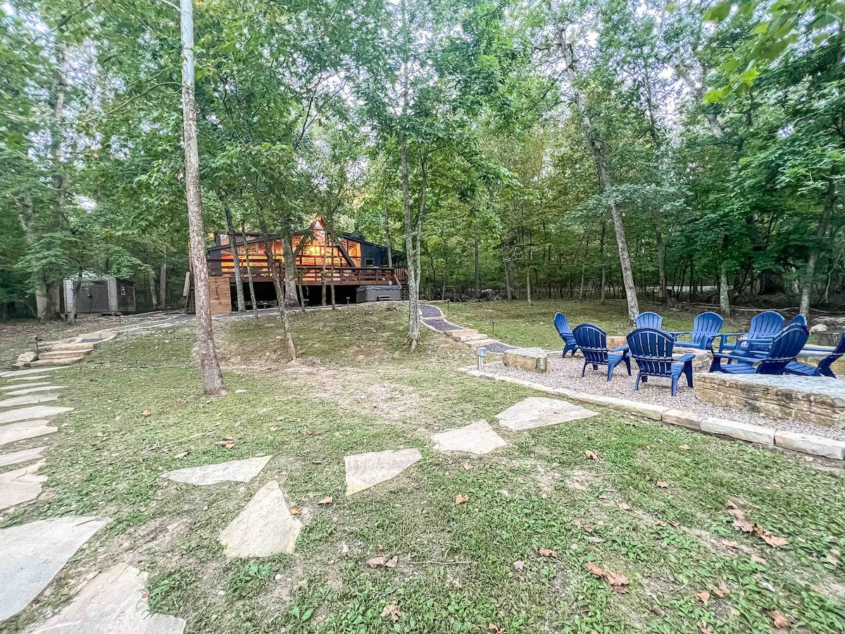 A serene outdoor space features a circular seating area with blue adirondack chairs surrounded by trees. A stone walkway leads to a chalet nestled among the woods, providing a cozy retreat with a warm glow emanating from its windows.