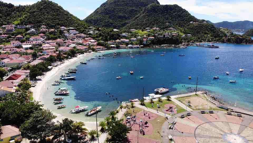 The image captures a coastal view of Terre-de-Haut, featuring a serene bay surrounded by lush hills. Various boats are seen moored in the clear water, while a sandy beach curves along the shore. A vibrant town with red-roofed buildings is positioned on the hillside.