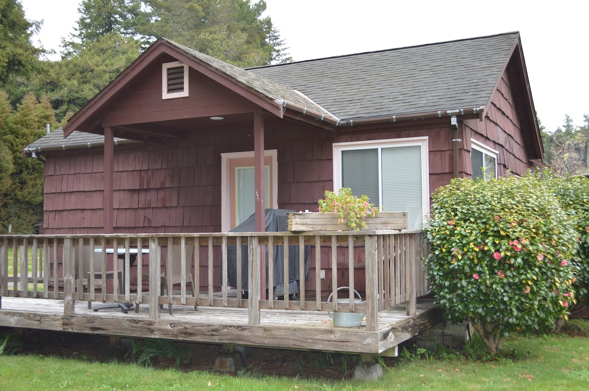 A rustic cabin with a red wooden exterior is featured, showcasing a spacious deck with a table and chairs. Lush greenery surrounds the cabin, and large windows allow natural light into the interior. The roof is peaked, adding to the charming cottage look.