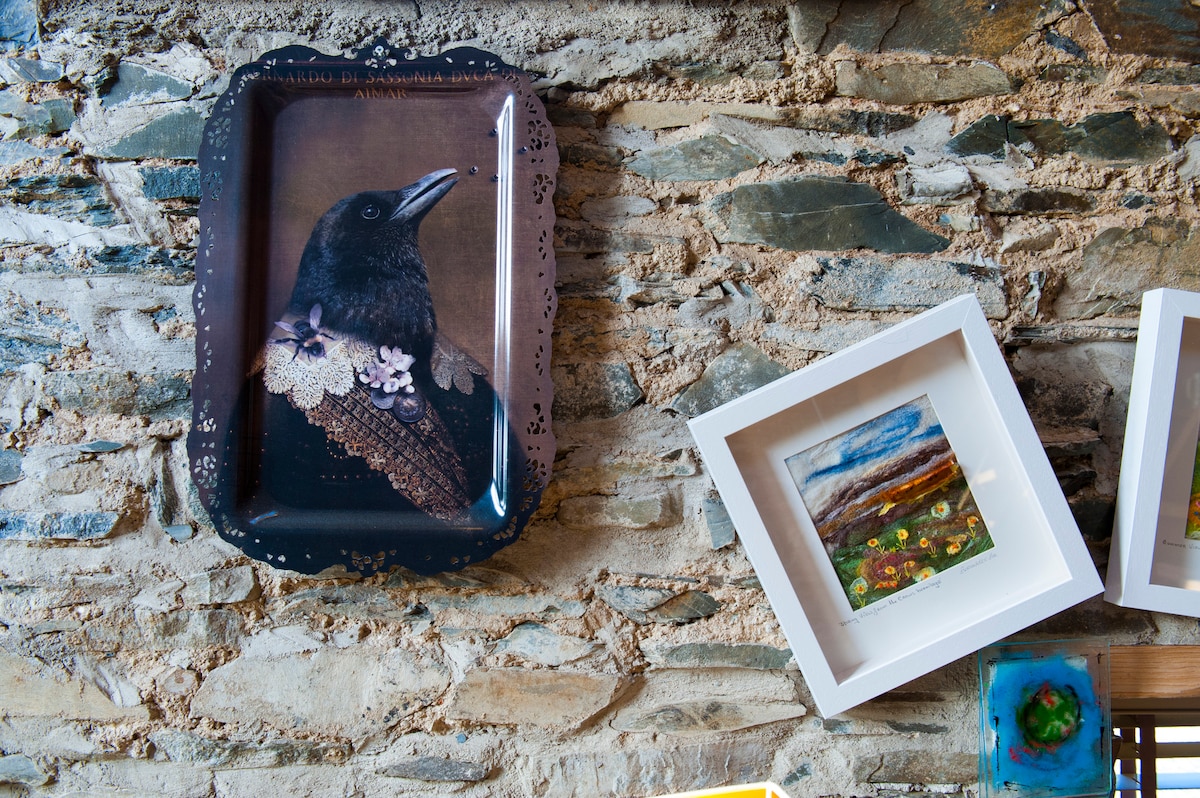 An exposed stone wall features a combination of artwork, including a dark tray adorned with a crow illustration and two framed pieces. One frame displays a colorful landscape, while the other portrays blooming flowers, adding character and artistic detail to the space.