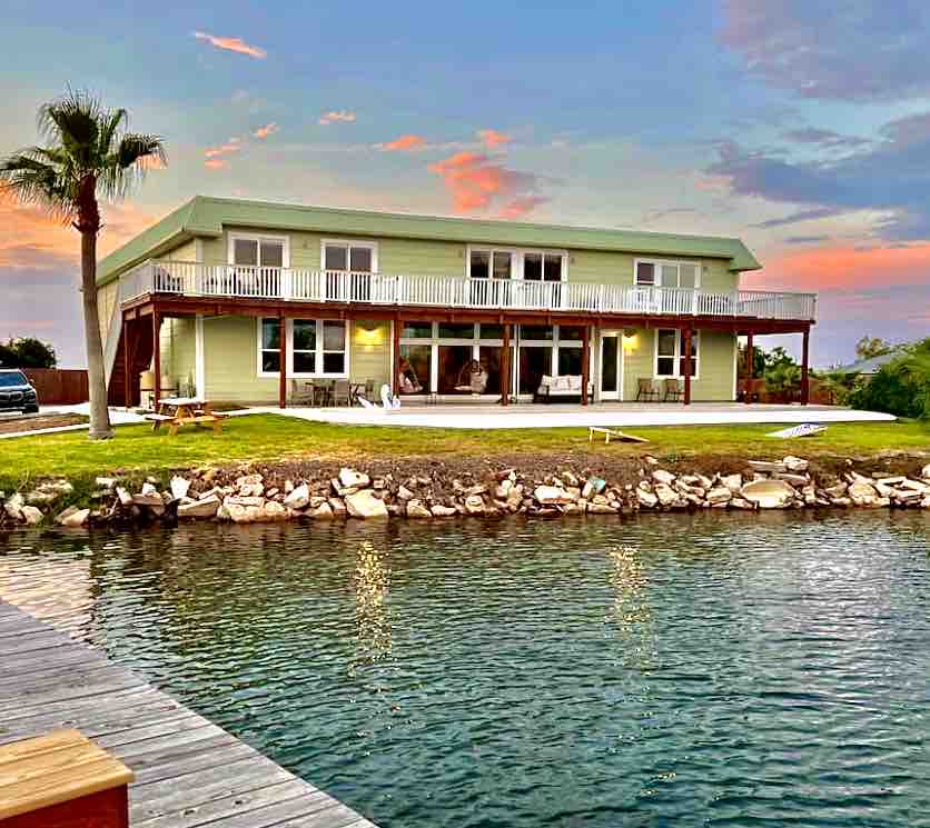 A two-story green house is positioned alongside a tranquil waterway, featuring a spacious balcony lined with white railings. The exterior is complemented by patio seating, and soft lighting reflects off the water at sunset, creating a serene outdoor ambiance.