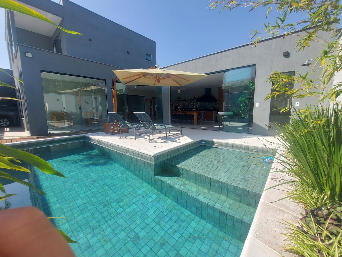 A modern outdoor space features a clear swimming pool with a shallow area, surrounded by lounge chairs and a large umbrella. The house's expansive glass doors provide views of the indoor dining area, illuminated by natural light.