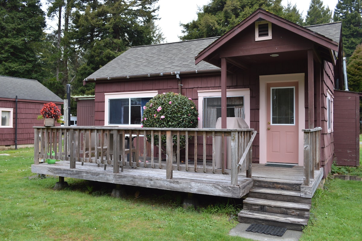 The cabin exterior features a rustic red wooden facade with a welcoming front porch. A wooden deck with a railing is highlighted by a potted plant and outdoor seating. Surrounding greenery complements the natural setting, contributing to a tranquil atmosphere.