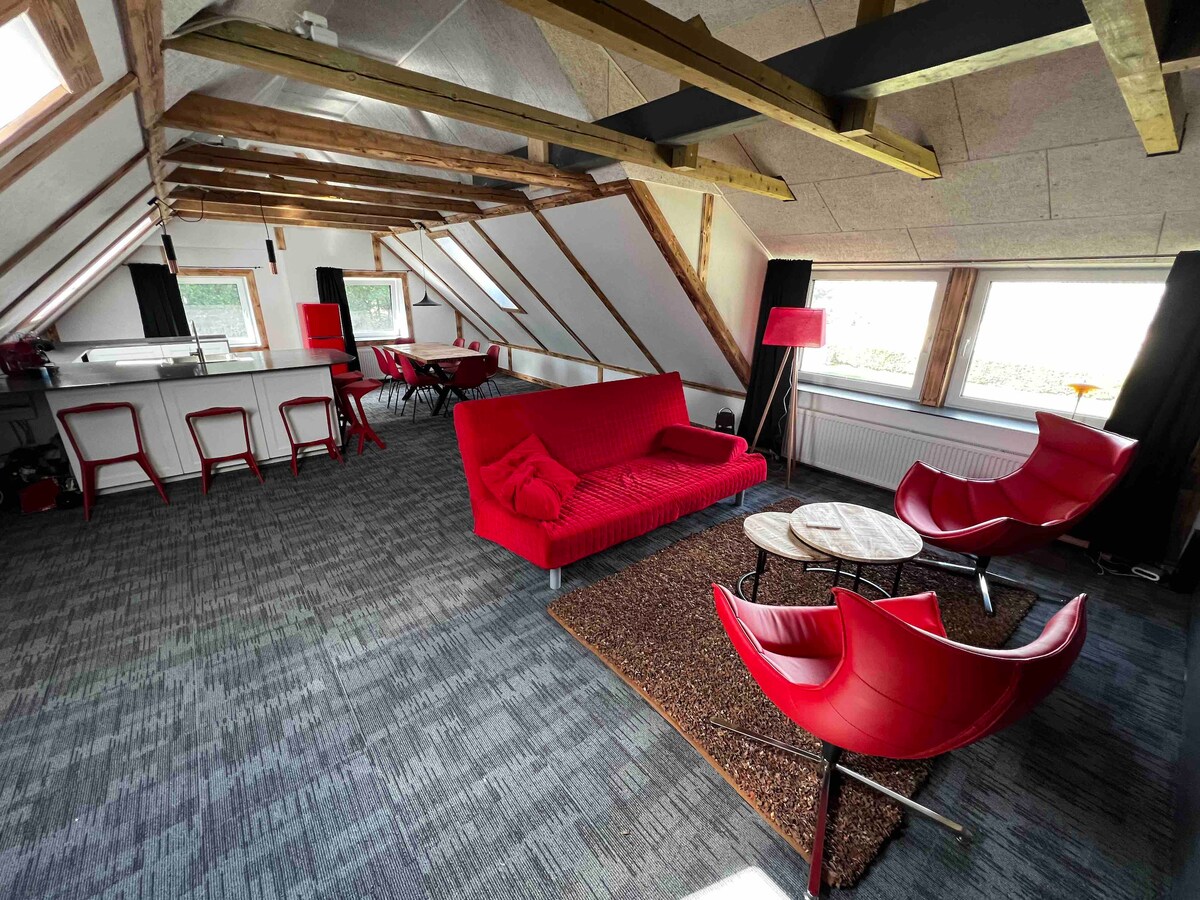 An open living area features a spacious red sofa complemented by two modern red chairs. Natural light enters through two large windows. A rustic coffee table is centered on a textured rug, while a dining area with several chairs is visible in the background.